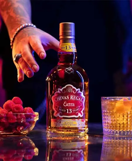 Foto de Chivas Extra 13 años x 700ml sobre mesa con coctel, fruta y una mano