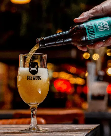 foto de Botella Brewdog Hazy x 330ml sirviendo una copa en una mesa de madera fondo oscuro con luces