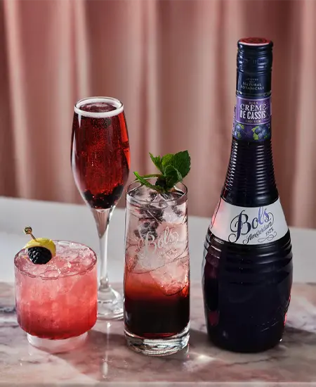 Mesa con 3 cocteles preparados con Bols Crema de Cassis x 700ml sobre mesa blanca y fondo rosado