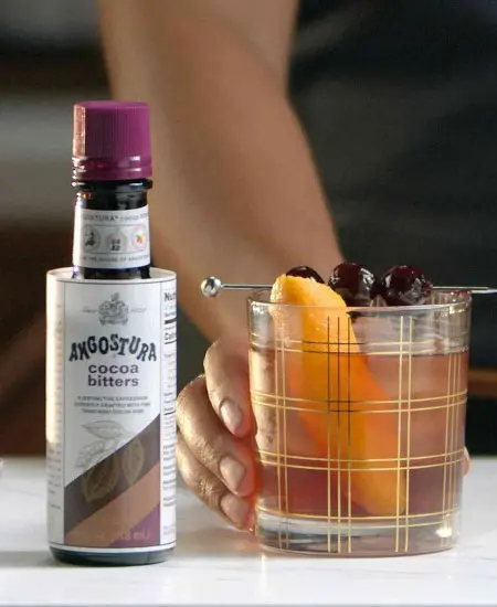 Angostura Cocoa (Esencias de gotas amargas) x 100ml junto a mano sosteniendo un coctel naranja en una mesa
